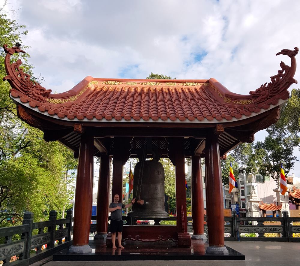 Dzwon świątynny Sajgon Mężczyzna w świątyni buddyjskiej w Sajgonie, stojący obok dzwonu ceremonialnego, na tle bogato zdobionej dzwonnicy.